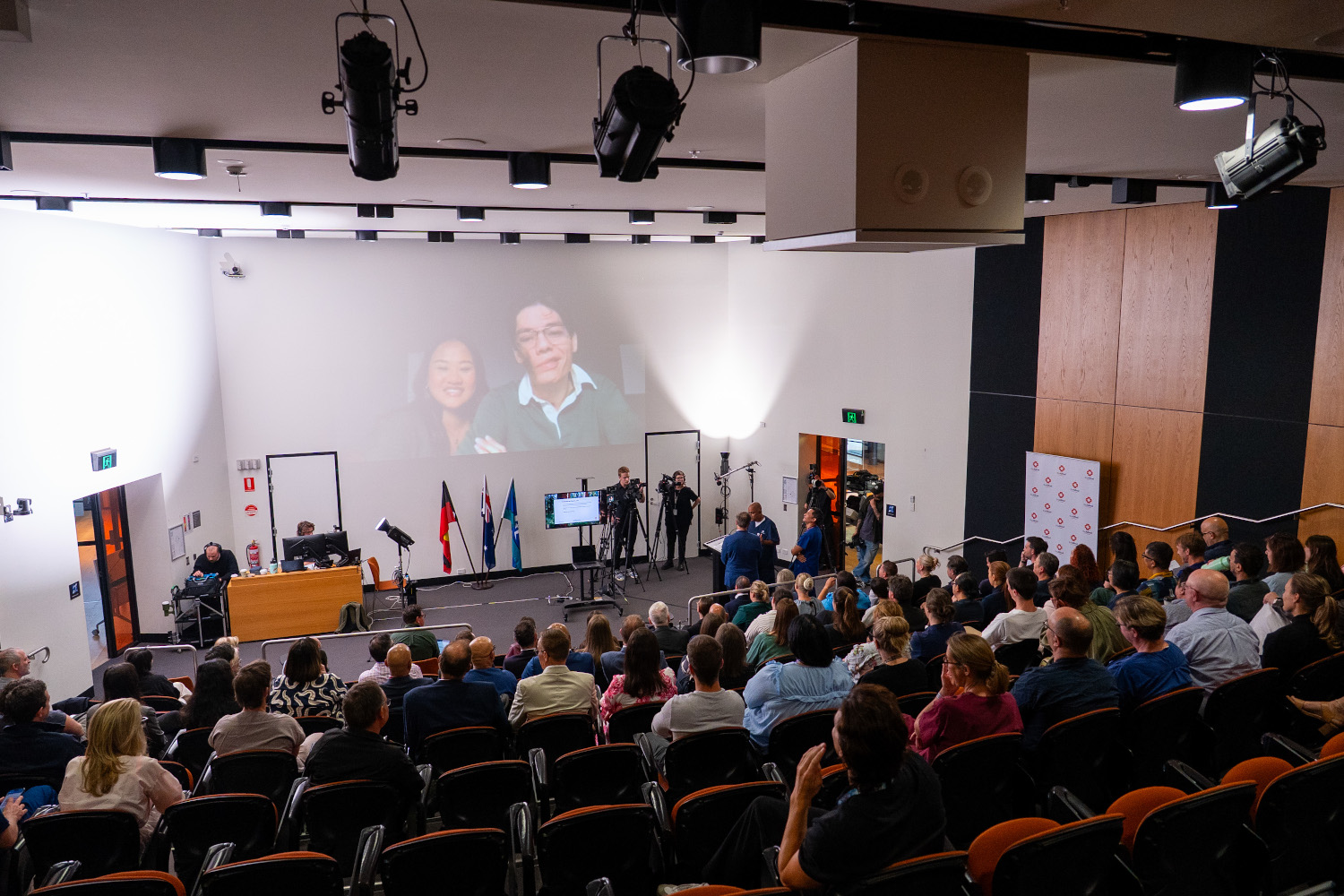 Travis and Jasmine on a projector screen facing a lecture theatre of clinicians, support and administrative staff