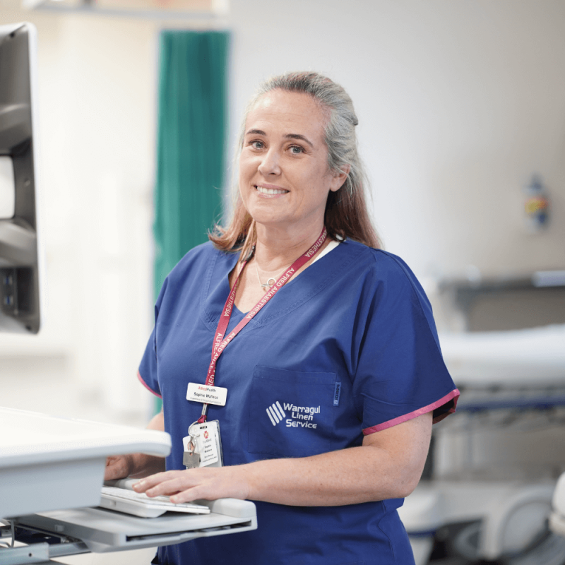Mature female nurse wearing blue scrubs and working at a wheeled monitor in a ward setting.