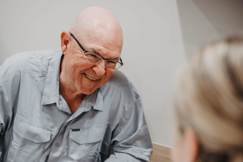 Older male clinical trials participant sitting with doctor. He is wearing a light blue buttoned shirt and has a bald head with glasses