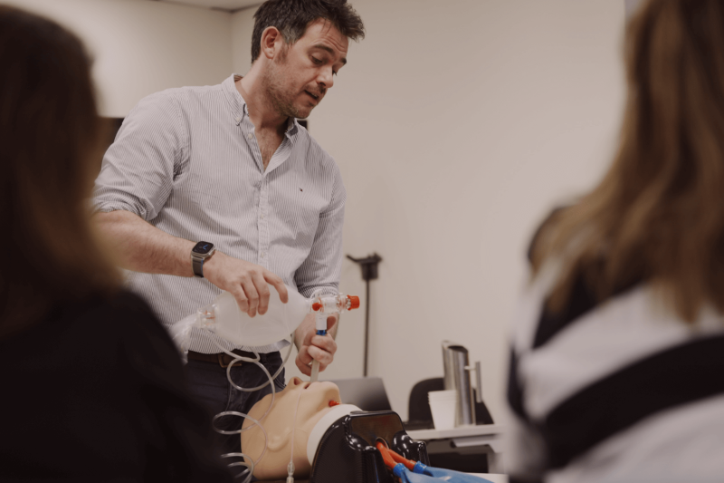 Man in collared shirt demonstrating airway equipment to two women in foreground of shot.